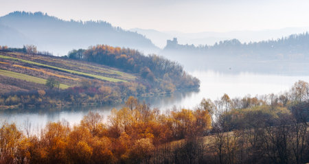 Autumn trees covered with yellow leaves illuminated by the sun. In the distance you can see the Pieniny Mountains and Nidzica Castle. This surface of water, a fisherman in a boatのeditorial素材