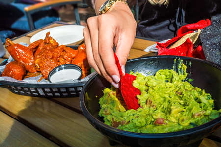 A girl dips into the guacamole with chips along with buffalo wings on the side as appetizers の写真素材