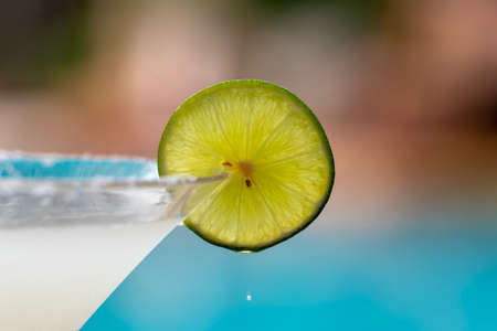 Closeup and focus on sliced lemon on glass of Margarita beside of swimming pool in blurred background with drop of lemonの写真素材
