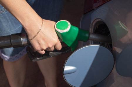 A woman in denim blue shorts with a fuelling nozzle pouring gasoline into the tank of the car at the...