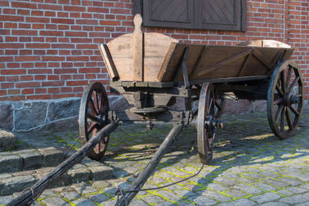A brown wooden vintage cart with wooden wheels, standing drawbar forward on an old gray stone sidewalk overgrown with grass against a red brick wall with a shuttered window at the stone porchの写真素材