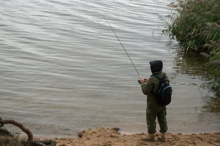 Fisherman standing on sandy shore of lake near a thicket of green reeds, holding fishing rod with...