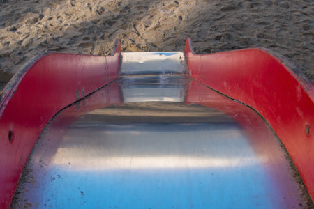 Slide with shiny smooth metal base and wooden red handrails descends steeply down to surface covered with sand with footprints on playground, top view. Concept: Prospect of falling from won positionsの写真素材