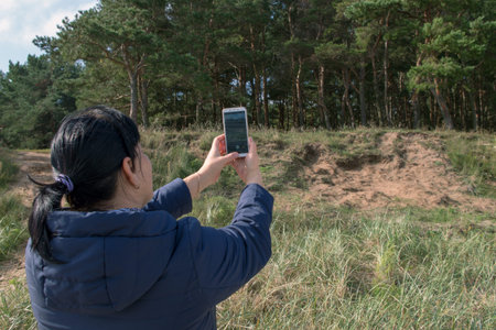 A woman with black hair with a ponytail in a blue hooded jacket taking a picture on a mobile phone...