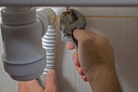 A man repairs plumbing under the sink with his right hand unscrews the nut fixing the flexible...