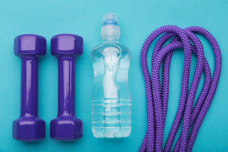 a bottle of water, lilac dumbbells and a skipping rope lined up in a row on a turquoise background,...
