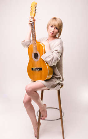 Young blonde lady in gray sweater sitting on chair and playing acoustic guitarの写真素材