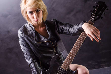 Beautiful young blonde dressed in black leather jacket with electric guitar on a black backgroundの写真素材