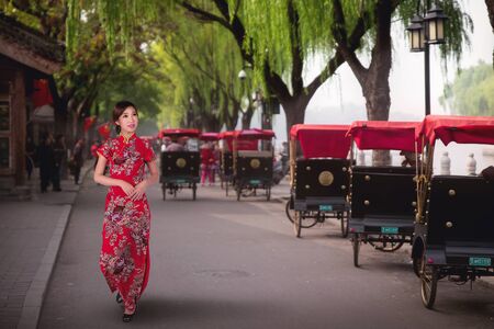 Chinese lady in red cheongsam dress walking near a Tourists riding Beijing traditional rickshaw in old China Hutongs in Beijing, China.の写真素材