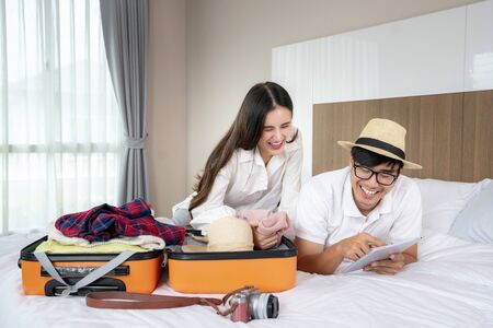 Happy asian couple packing suitcase on the bed in room use tablet for search travel trip onlineの写真素材