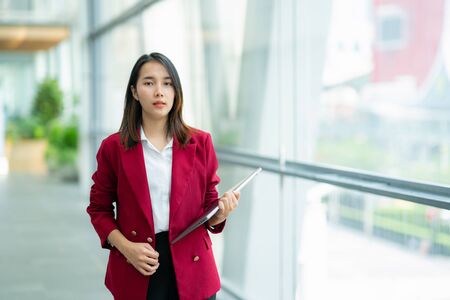 Beautiful business woman with long red hair walking near office building. Young smiling woman goes with laptop overlooking the city center.の写真素材