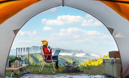 Woman working drink a coffee and working with computer note book between tent camping with summer and mountain background on morning timeの写真素材