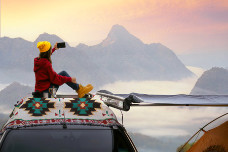 Traveller woman sit on the roof top tarp and car and drink a coffee with morning sunrise and fog on the mountain view, Chiangmai, Thailandの写真素材