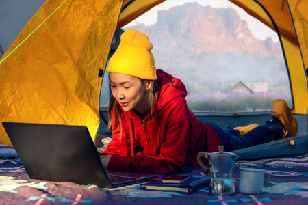 Woman working drink a coffee and working with computer note book between tent camping with summer and mountain background on morning timeの写真素材