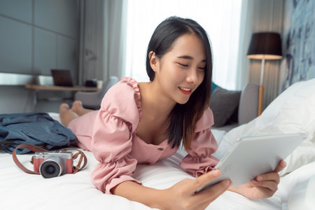 Asian traveller woman take travel bag sleep on the bed and check masage in her mobile in her hotel room after finish her travel tripの写真素材