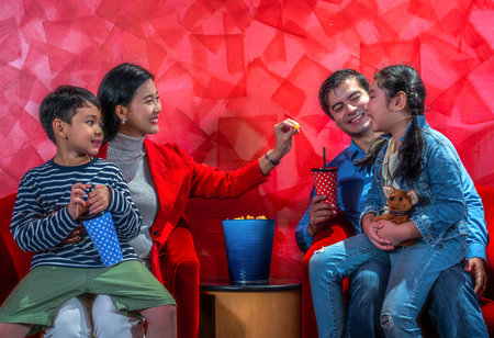 Happy Indonesian family waiting movie in front of cinema with popcorn and beverage on the sofa seat in weekend holidayの写真素材