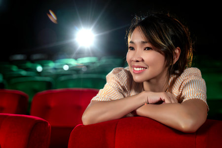 Singgle middle age asian woman looking to movie screen in cinema theater with red seat and light from a projector machine backgroundの写真素材