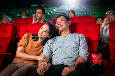 Happy trip of asian couple sweet sit togather on red seat and looking  movie in cinema with fun a popcorn on hand, people relax and holiday activity on a weekendの写真素材