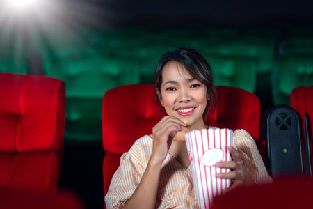 Singgle middle age asian woman looking to movie screen in cinema theater with red seat and light from a projector machine background with popcornの写真素材