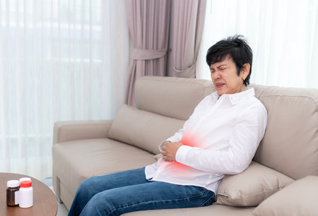 Stressed mature senior woman stomachache at sofa in living room, suffering from headache, migraine, blood pressure problem, health problems, feeling strong painの写真素材