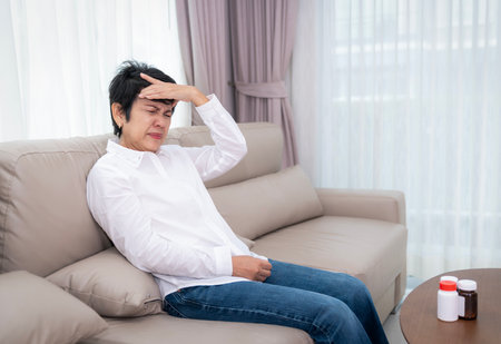 Stressed mature senior woman touching head with closed eyes at sofa in living room, suffering from headache, migraine, blood pressure problem, health problems, feeling strong painの写真素材