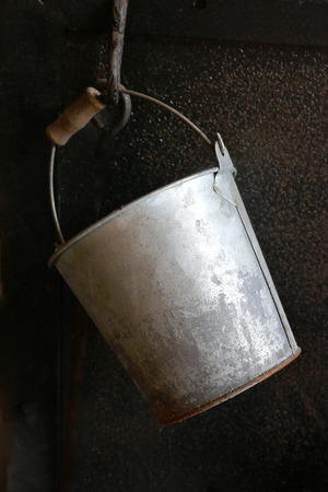 Old weathered rusty metal pail with a wooden handle hanging in front of black background. Closeの写真素材