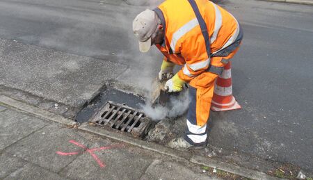 Road construction, road rehabilitation, asphalting. A worker in a protective suit repairs damage to a road by pouring boiling hot asphalt into the pothole next to the drain.の写真素材