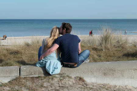 Scharbeutz, Germany - February 25, 2021: Young couple sits on a wall and looks over the dune to the beach and the sea in Scharbeutz, a seaside resort on the Baltic Sea coast in Schleswig-Holsteinのeditorial素材