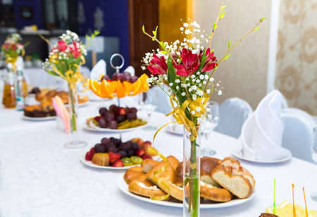 Beautiful table setting with a bouquet of flowers. White tablecloth with white napkins. Bright fruits and vegetables on the table. Wedding table. Holiday, Christmas, new year, birthday. Blurred backgroundの写真素材