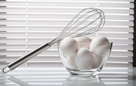 Still life of metal whisk on bowl full of raw eggs on reflective kitchen table over white jalousie.の写真素材