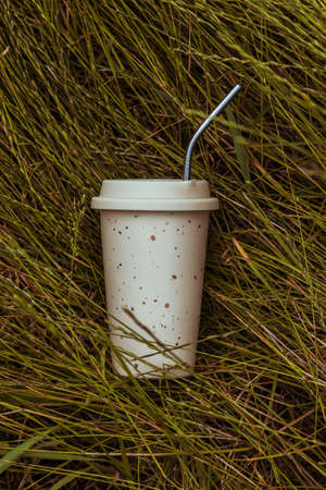 A beige reusable ceramic drink cup and a reusable straw lie on the green grass.の写真素材