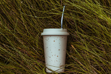 A beige reusable ceramic drink cup and a reusable straw lie on the green grass. Horizontal photo.の写真素材