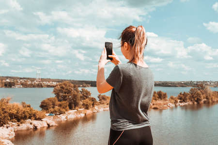 A girl with her hair pulled back stands on the edge of a cliff and takes a picture of the scenic landscape with her phone.の写真素材