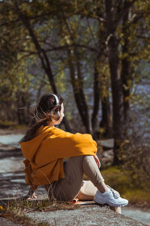 A girl in a yellow sweater is sitting on the edge, listening to music with white headphones and looking at the river. Vertical photo.の写真素材