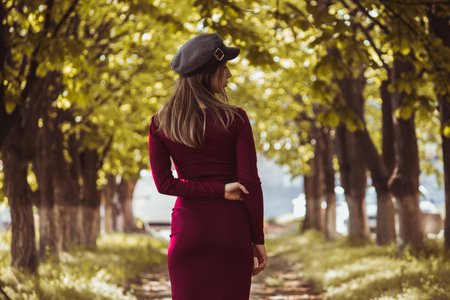 A slender girl in a burgundy dress and a headdress turned her back against the background of an arch of trees on a autumn sunny day.の写真素材