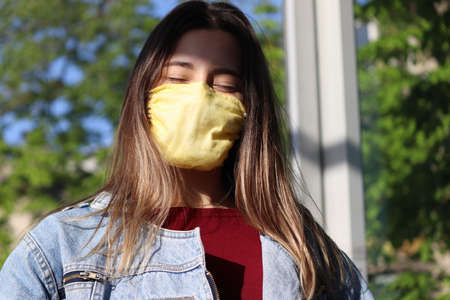 A girl with long hair in a yellow mask closes her eyes because of the bright sun at the bus stop.の写真素材