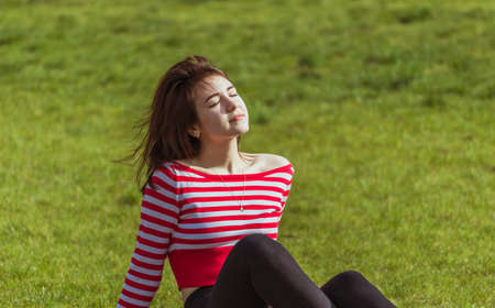 A girl with dark hair sits on the grass with closed eyes and enjoys the sun's rays.の写真素材