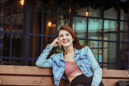 A satisfied smiling girl sits on a bench near a cafe and looks up.の写真素材