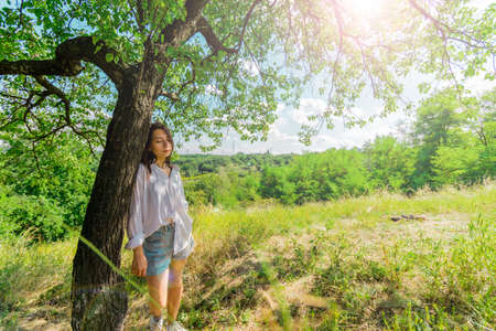 A girl with dark hair and a white shirt is leaning against a tree against the background of a meadow. Relaxation and rest from the bustle of the city. High quality photoの写真素材