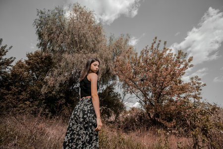 A beautiful slender girl in a long skirt stands against the background of autumn dry trees and a gray sky. Autumn mood and melancholy. High quality photoの写真素材