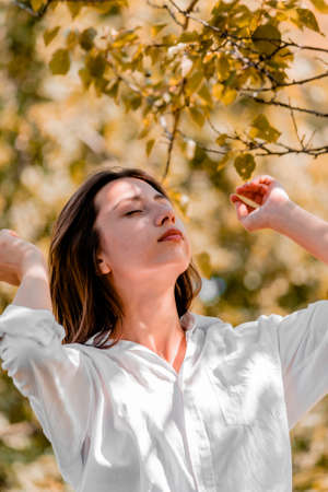 Random photo. A beautiful girl in a white shirt straightened her hair with her hands against the backdrop of trees on a sunny day. High quality photoの写真素材