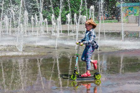 Ukraine - May 26, 2018. A girl rides a scooter in a puddle next to the fountain on a sunny day. Dnieper embankment, Ukraine. High quality photoのeditorial素材