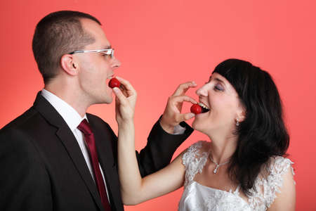 Happy smiling bride and groom young happy couple playfully eating strawbery on red backgroundの写真素材
