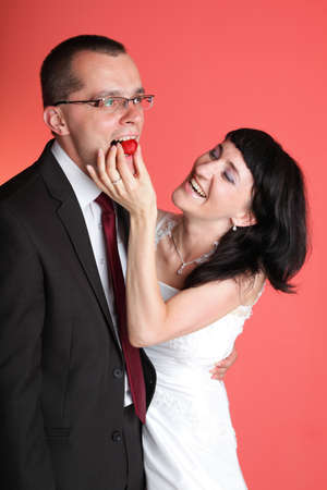 Happy smiling bride and groom young happy couple playfully eating strawbery on red backgroundの写真素材