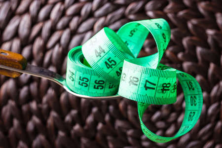 Diet food healthy lifestyle and slim body concept. Closeup green measuring tape centimeter on kitchen spoon brown backgroundの写真素材