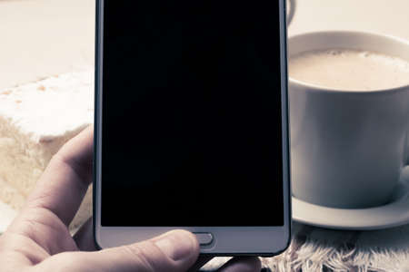 Closeup hand with smartphone black screen and cup of coffee and cake in the backgroundの写真素材