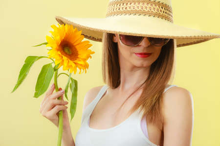 Closeup of attractive summer woman in sunglasses and straw hat with sunflower in hand on yellow backgroundの写真素材