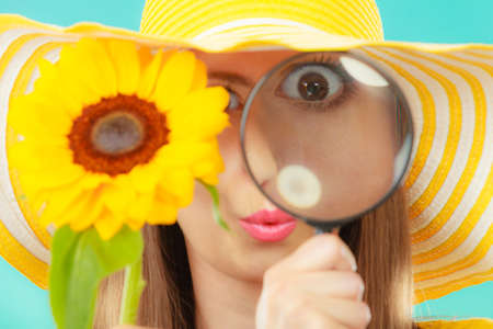Botanist woman funny face expression in yellow hat examining flower looking through magnifying glass on blue backgroundの写真素材
