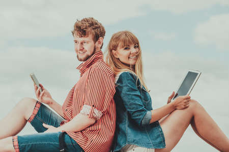 Modern technology devices. Young couple outdoor against sky man with smartphone, woman with tablet enjoying summer holidayの写真素材