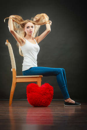 Broken heart love concept. Sad melancholy woman sitting on chair red heart pillow on floor dark backgroundの写真素材